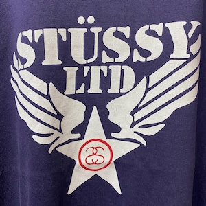 May include: A close-up of a navy blue sweatshirt with a white graphic. The graphic features the text "ST&Uuml;SSY LTD" above a star with a red and white logo, and white wings extending from the star.