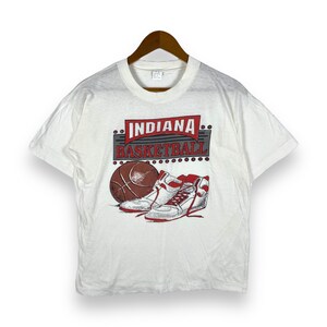 Pode incluir: Camiseta branca com um gráfico vintage de uma bola de basquete, tênis e o texto "INDIANA BASKETBALL".