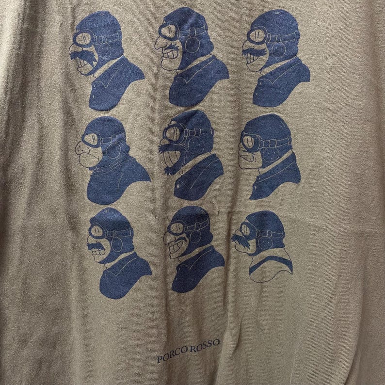 May include: A tan t-shirt featuring a repeating pattern of a cartoon aviator's head in navy blue. The aviator has goggles and a mustache. The text "PORCO ROSSO" is printed in white at the bottom of the shirt.