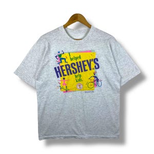 May include: A light gray t-shirt with a graphic featuring the Hershey's logo in purple and the words "helped" and "help kids." The graphic includes illustrations of a skateboarder, roller skater, and cyclist on a yellow background.