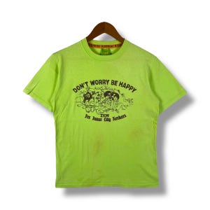 以下が含まれることがあります： ライムグリーンのTシャツに、黒いグラフィックプリントが施されています。プリントには「Don't Worry Be Happy」という文字と、丘に座っている人々の漫画が描かれています。「ZION For Inner City Rockers」という文字が、グラフィックの下にプリントされています。