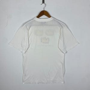 May include: White t-shirt with a graphic of three yellow crowns on the front. The graphic is a simple line drawing with a black outline.