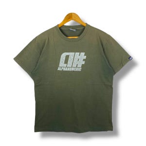 Könnte beinhalten: Olivgrünes T-Shirt mit dem Text "ALPHANUMERIC" in weißer, blockiger Schrift. Das Shirt ist kurzärmlig und hat einen Rundhalsausschnitt. Das Shirt hängt an einem Holzbügel.
