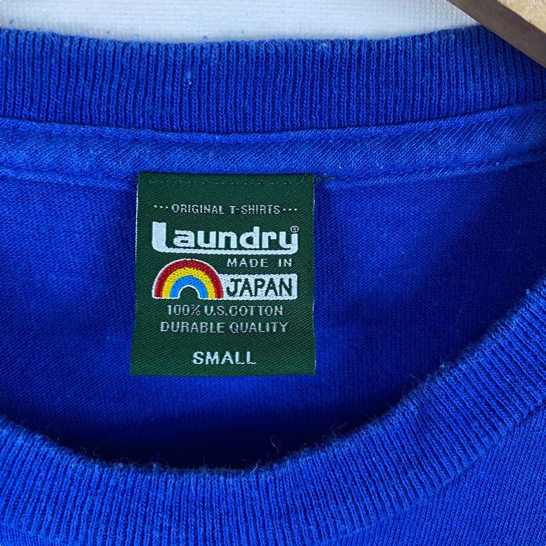 May include: A blue t-shirt with a green tag that says "Laundry" in white letters with a rainbow underneath. The tag also says "Made in Japan", "100% U.S. Cotton Durable Quality", and "Small".
