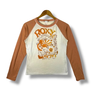 May include: A long-sleeved raglan shirt with a white body and rust-coloured sleeves. The shirt features a retro graphic with the word "ROXY" and the number "1970" in orange, along with floral and sun designs.