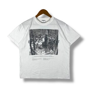 以下が含まれることがあります： 白地に黒と白のグラフィックプリントが施されたTシャツ。水の中に立っている女性が描かれている。Tシャツには「The Commander」と「Eyes Open」の文字がプリントされている。