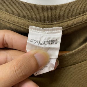 May include: Close-up of a brown t-shirt with a white tag. The tag has Japanese text printed on it. The t-shirt is made of a soft, textured fabric. The tag is held by a person's hand.