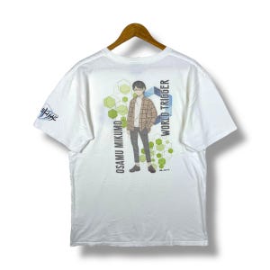 May include: White t-shirt featuring a graphic of Osamu Mikumo from World Trigger. The design includes a character illustration, text, and geometric shapes in green and blue. The shirt is hanging on a wooden hanger.