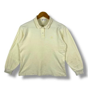 May include: Cream-colored long-sleeve polo shirt with a collar and three buttons. The shirt has a small logo on the chest and ribbed cuffs. The shirt is hanging on a wooden hanger.