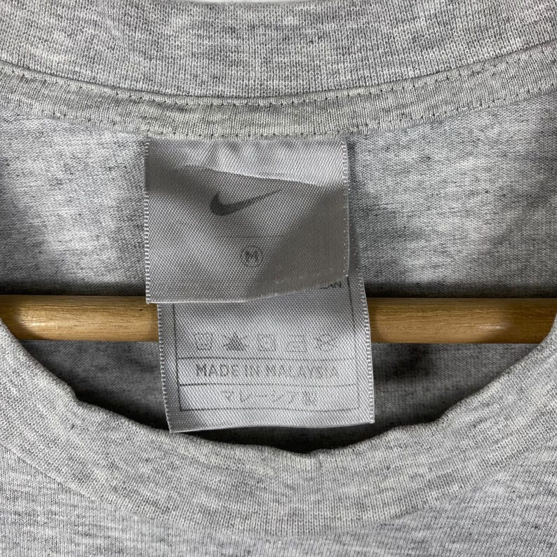 Może przedstawiać: Szary T-shirt z białą metką z napisem "Nike" i "Wyprodukowano w Malezji".