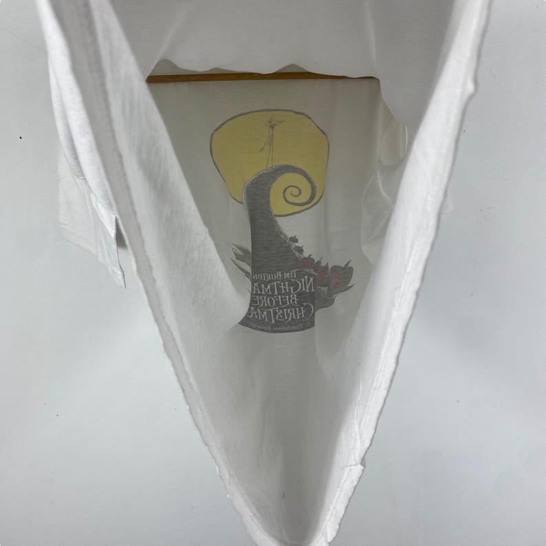 Puede incluir: Camiseta blanca con un estampado gr&aacute;fico de Jack Skellington de la pel&iacute;cula "Pesadilla antes de Navidad". El gr&aacute;fico muestra a Jack Skellington de pie en una colina con una luna llena detr&aacute;s de &eacute;l. El texto "Pesadilla antes de Navidad" est&aacute; impreso debajo del gr&aacute;fico.