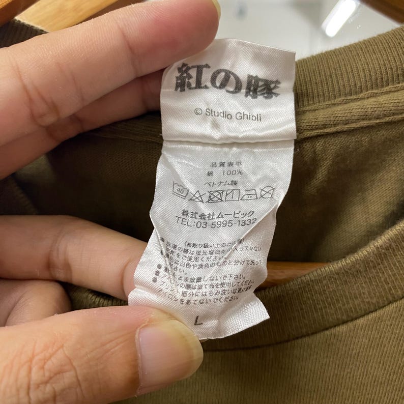 May include: Close-up of a tag from a brown t-shirt. The tag features Japanese text, the Studio Ghibli logo, and washing instructions. The tag also includes the text "100%" and "Made in Vietnam". The size is labeled as "L".