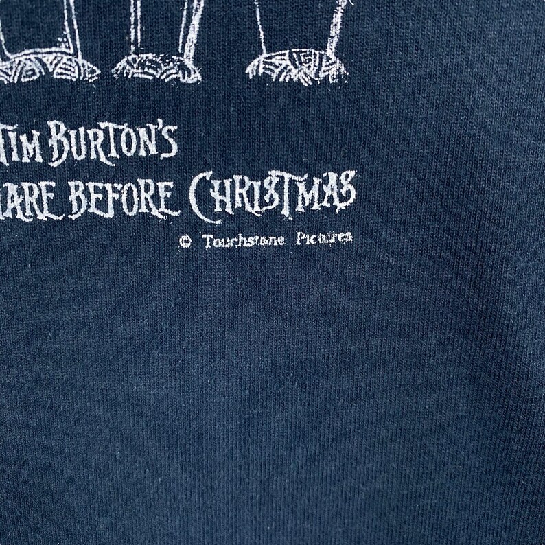 Puede incluir: Tela azul marino oscuro con texto blanco que dice "Tim Burton's The Nightmare Before Christmas" y "&copy; Touchstone Pictures". El texto est&aacute; en una fuente estilizada. La tela parece ser una camiseta u otra prenda similar.