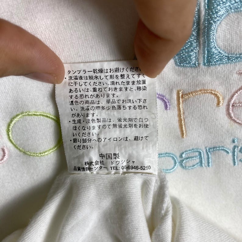 May include: White t-shirt with a colorful embroidered logo that says "Paris". The care instructions on the tag say to avoid tumble drying, wash dark colors separately, and use non-fluorescent detergent for light colors. The tag also says to avoid ironing the decorative parts of the shirt.