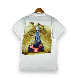 May include: White t-shirt with a graphic of a gray alien mowing a lawn with a red lawnmower. The alien is wearing blue overalls and a white shirt. The lawn is green and there are small gray aliens in the background.