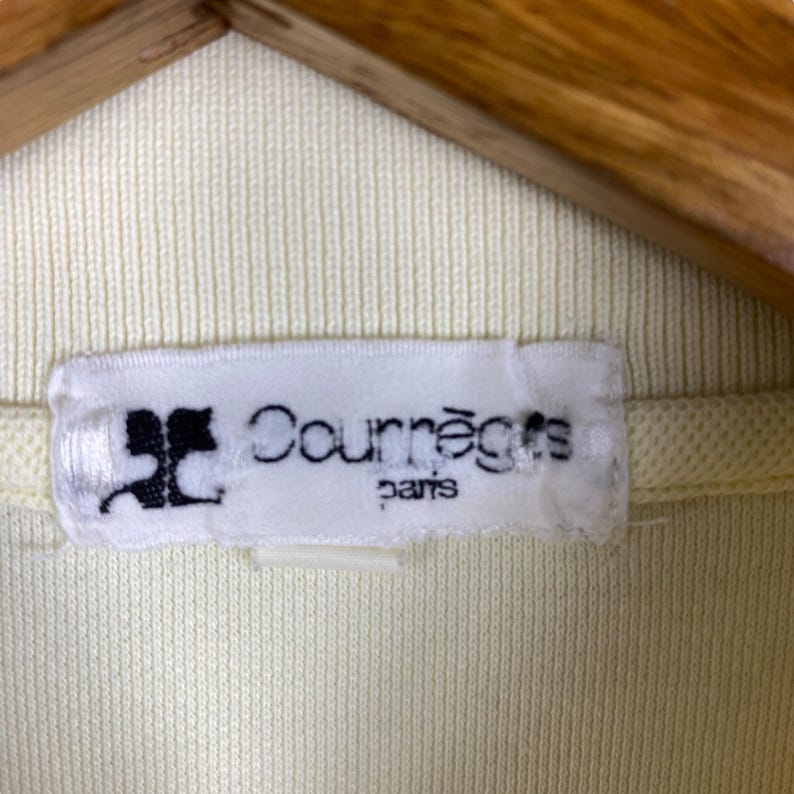 May include: Close-up of a pale yellow garment with a white label. The label features the brand name "Courr&egrave;ges" in black text, with "paris" below. The garment is hanging on a wooden hanger.