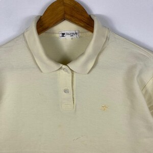 May include: Cream-colored polo shirt with a classic collar and short sleeves. The shirt features a three-button placket and a small embroidered logo on the chest. The Courr&egrave;ges Paris label is visible on the collar.