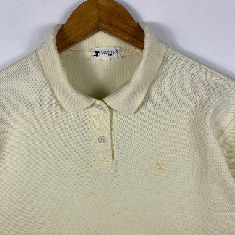 May include: Cream-colored polo shirt with a classic collar and short sleeves. The shirt features a three-button placket and a small embroidered logo on the chest. The Courr&egrave;ges Paris label is visible on the collar.