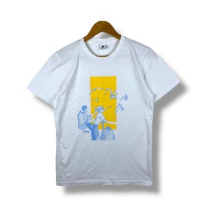 Könnte beinhalten: Weißes T-Shirt mit einem gelben rechteckigen Grafikdesign. Die Grafik zeigt eine blaue Illustration von zwei Personen und fliegenden Vögeln. Das T-Shirt hängt an einem Holzkleiderbügel.