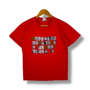 May include: A red t-shirt featuring a graphic of multiple faces and the word "WORST" in green. The t-shirt is displayed on a wooden hanger. The shirt has short sleeves and is set against a white backdrop.