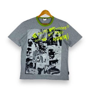 May include: Gray short-sleeved t-shirt with a green trim around the neck. The shirt features a collage of black and white images with the text "If you're not outraged you're not paying attention!" in neon green.