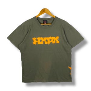 Peut inclure: T-shirt vert olive avec le mot "ITPX" en orange vieilli sur la poitrine. Le t-shirt est suspendu à un cintre en bois sur fond blanc. Un petit motif orange est visible en bas à droite.
