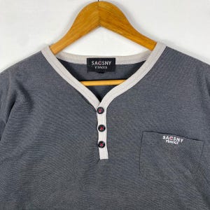 K&ouml;nnte beinhalten: Ein graues, gestreiftes Langarmshirt mit wei&szlig;em Besatz am Ausschnitt und einer kleinen Tasche auf der linken Seite. Das Shirt hat eine Knopfleiste und der Markenname "SACOSNY Y'SACCS" ist auf dem Shirt aufgedruckt.