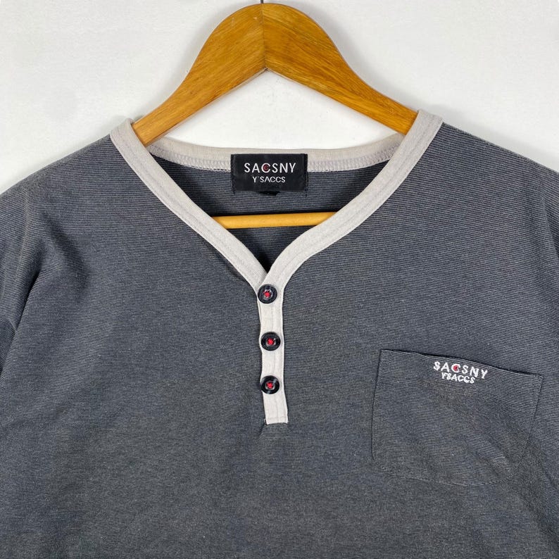 K&ouml;nnte beinhalten: Ein graues, gestreiftes Langarmshirt mit wei&szlig;em Besatz am Ausschnitt und einer kleinen Tasche auf der linken Seite. Das Shirt hat eine Knopfleiste und der Markenname "SACOSNY Y'SACCS" ist auf dem Shirt aufgedruckt.