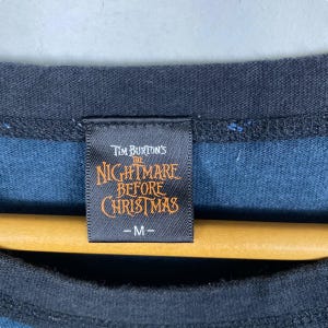 Puede incluir: Primer plano de una etiqueta de camiseta negra y azul con el texto "Tim Burton's The Nightmare Before Christmas" en naranja y blanco. La etiqueta tambi&eacute;n tiene la talla "-M-" impresa. La camiseta est&aacute; colgada en una percha de madera.