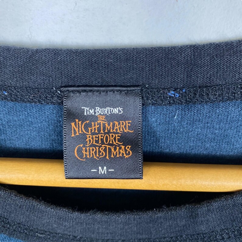 Puede incluir: Primer plano de una etiqueta de camiseta negra y azul con el texto "Tim Burton's The Nightmare Before Christmas" en naranja y blanco. La etiqueta tambi&eacute;n tiene la talla "-M-" impresa. La camiseta est&aacute; colgada en una percha de madera.