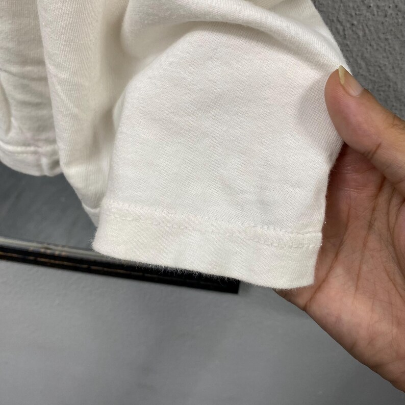 May include: A plain white long-sleeve shirt featuring a simple crew neckline and a straight hemline with double-stitched detailing along the cuff. The fabric has a soft, matte texture and a clean, solid white color throughout the garment.