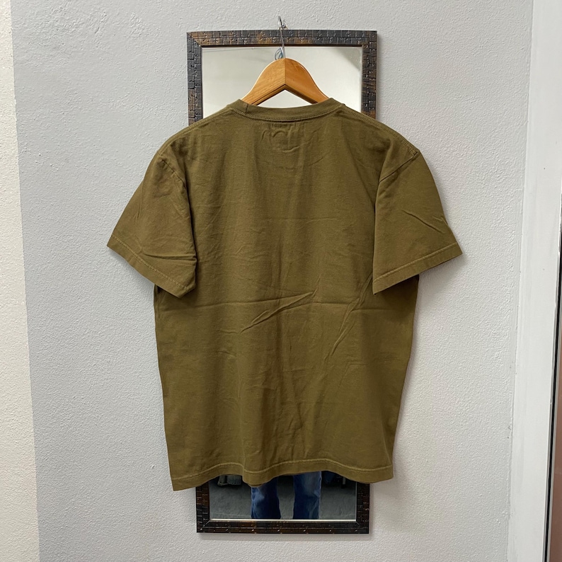 May include: Olive green short-sleeve t-shirt hanging on a wooden hanger. The shirt is displayed against a neutral wall with a framed mirror. The back of the shirt is visible, showing the neckline and the texture of the fabric.