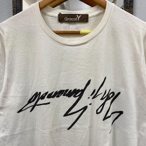 May include: A white short-sleeved t-shirt with a crew neckline. The chest features a large, black, stylized handwritten graphic print. A brown rectangular label at the inner neckline displays the text Ground Y in white lettering.