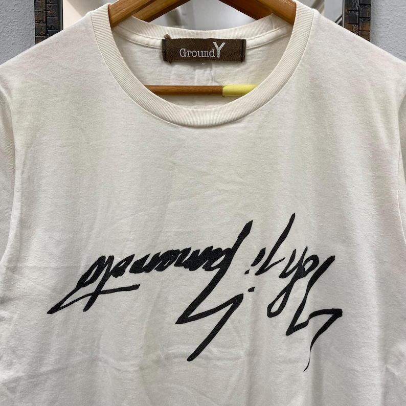 May include: A white short-sleeved t-shirt with a crew neckline. The chest features a large, black, stylized handwritten graphic print. A brown rectangular label at the inner neckline displays the text Ground Y in white lettering.
