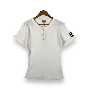 May include: White short-sleeved shirt with a button-up collar and a small logo patch on the sleeve. The shirt is made of a ribbed fabric and has a relaxed fit.