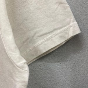 May include: A close-up view of a short sleeve on a plain off-white t-shirt. The fabric shows a visible woven texture with a double-stitched hem along the edge of the sleeve. The garment is made of a light-colored cotton material with clean seams.