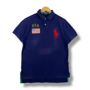 Peut inclure: Un polo bleu marine avec un logo de joueur de polo brodé en rouge et un petit drapeau américain avec le mot "USA" au-dessus.