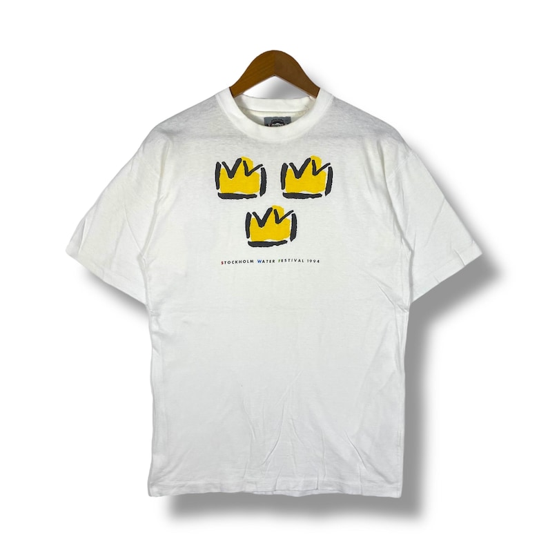 May include: White t-shirt with a graphic of three yellow crowns and the text "STOCKHOLM WATER FESTIVAL 1994".