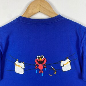 May include: Blue t-shirt featuring a red Sesame Street character hanging from a clothesline with the word "Laundry" on a white shirt.