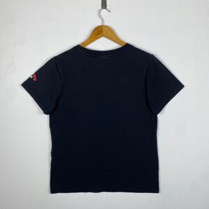 Pode incluir: T-shirt preto pendurado em um cabide de madeira. A camisa de manga curta tem gola redonda e um pequeno gr&aacute;fico vermelho na manga esquerda. A camisa &eacute; feita de um material macio.