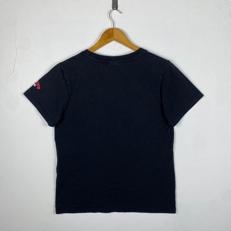 Pode incluir: T-shirt preto pendurado em um cabide de madeira. A camisa de manga curta tem gola redonda e um pequeno gr&aacute;fico vermelho na manga esquerda. A camisa &eacute; feita de um material macio.