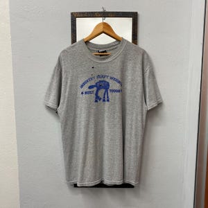 May include: A heather gray t-shirt with a blue graphic that reads "INDUSTRY HEAVY WEIGHT BUILT TOUGH FUCT". The shirt is hanging on a wooden hanger against a wall with a mirror.