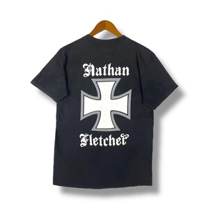 May include: Black t-shirt with a white iron cross and the text "Nathan Fletcher" printed on the back.
