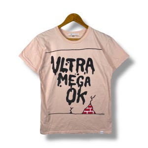 Puede incluir: Camiseta rosa claro con la frase "ULTRA MEGA OK" en negro con tipografía goteante. Debajo, un dibujo de un muro de ladrillos. La camiseta está colgada en una percha de madera.