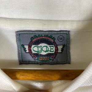May include: White polo shirt with a gray and green embroidered logo that says "C.I.C.E" and "Team Spirit". The logo is surrounded by a wreath of green leaves. The shirt has a size tag that says "M".
