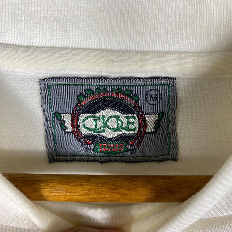 May include: White polo shirt with a gray and green embroidered logo that says "C.I.C.E" and "Team Spirit". The logo is surrounded by a wreath of green leaves. The shirt has a size tag that says "M".
