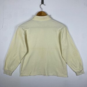 May include: Cream-colored long-sleeve shirt with a mock neck collar. The shirt is hanging on a wooden hanger against a plain white wall. The sleeves have a slight puff at the shoulders and end in ribbed cuffs.