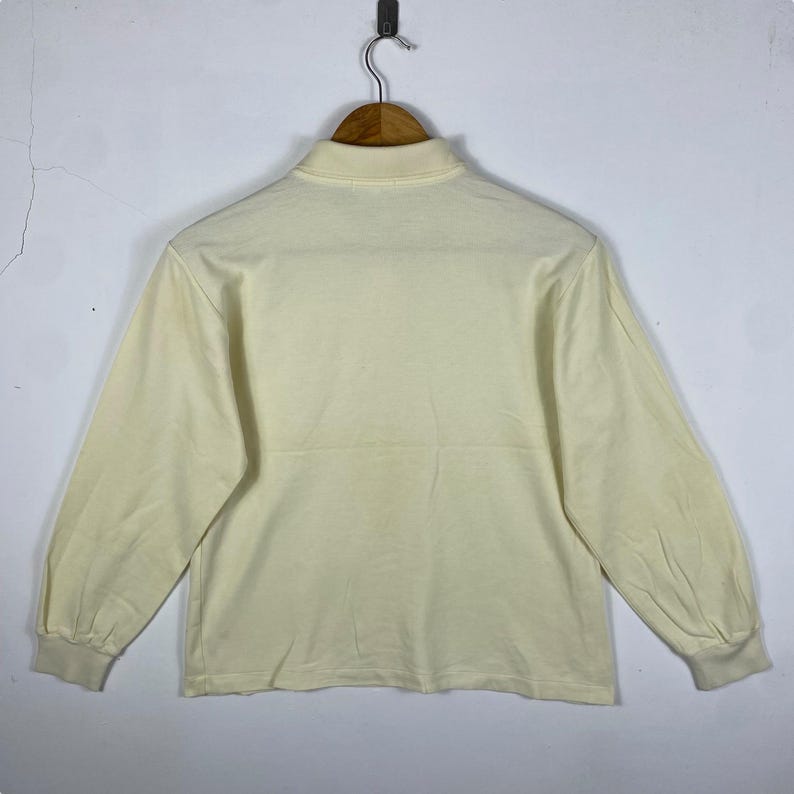 May include: Cream-colored long-sleeve shirt with a mock neck collar. The shirt is hanging on a wooden hanger against a plain white wall. The sleeves have a slight puff at the shoulders and end in ribbed cuffs.