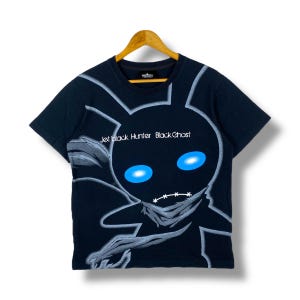 May include: Black t-shirt with a graphic of a black cartoon character with blue eyes and the text "Jet Black Hunter Black Ghost". The shirt is hanging on a wooden hanger.