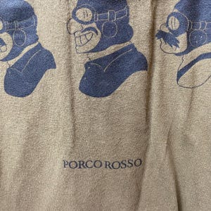 May include: Close-up of a tan t-shirt featuring a repeating blue graphic of a character wearing goggles and a pilot's cap. The text "PORCO ROSSO" is printed in blue at the bottom of the shirt.